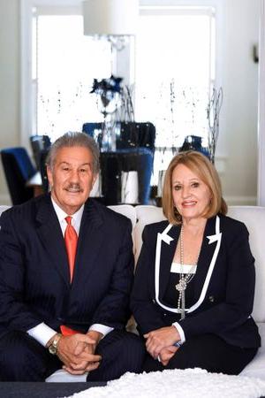 a photograph of Gigi Manchin and Mark Manchin sitting side by side on a white couch wearing formal clothing.  They are smiling at the camera.  In the background is bright white light shining in from outside and a long table with chairs around it